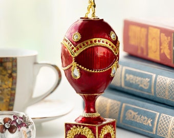 以下が含まれることがあります： 金色の置物が乗った赤と金のファベルジェの卵のレプリカ。卵は金色のアクセントがついた正方形の台座の上に置かれています。