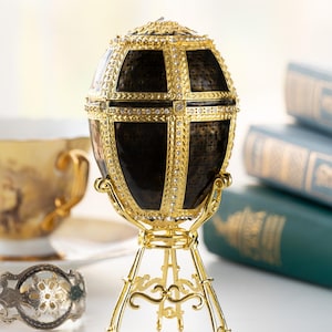 May include: A gold and black jeweled egg on a gold stand. The egg is decorated with a geometric pattern and has a shiny finish.