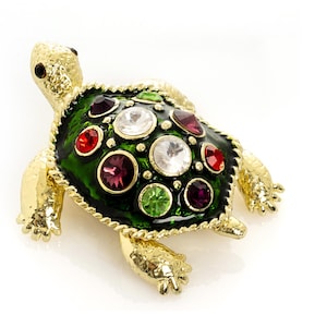 May include: A gold-toned turtle brooch with a green shell adorned with colorful gemstones. The brooch features a detailed design with red, purple, green, and clear stones, creating a sparkling effect. The turtle's head and legs are also gold-toned.