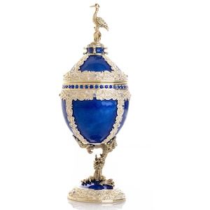 May include: A cobalt blue decorative egg-shaped object with gold filigree accents and a small bird figurine on top. The egg is supported by a gold-colored, ornate stand. Small blue gemstones are set into the gold trim.
