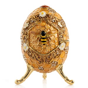 Könnte beinhalten: Dekorative, eiförmige Schmuckschatulle mit einem goldenen Bienenmotiv. Die Schatulle ist überwiegend goldfarben, verziert mit einem Wabenmuster, weißen floralen Details und einem roten Edelstein. Sie steht auf drei goldenen Beinen.
