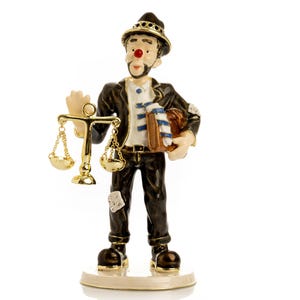 May include: A whimsical figurine of a man in a black suit, holding a golden scale in one hand and a brown satchel with a blue and white striped item in the other. The figure has a red nose, a small black hat, and a white shirt. The figurine stands on a beige base.