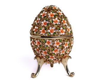 May include: A gold egg-shaped trinket box with a floral design. The box is decorated with orange and white flowers and green leaves. The box has three legs and is open.