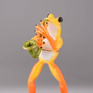 May include: A yellow and orange frog figurine playing a green saxophone, standing on a green leaf with a gold trim.