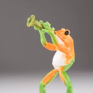 May include: A green and orange frog figurine playing a trumpet, standing on a green leaf base. The frog has black eyes and a white belly.