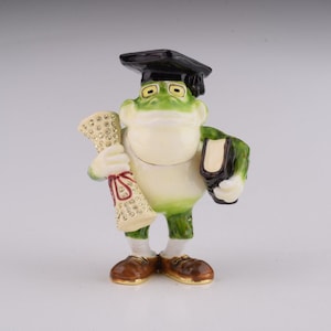 May include: A green frog figurine wearing a black graduation cap and holding a scroll and a book. The frog is wearing brown shoes and has a white belly.