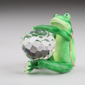 May include: A green frog figurine holding a clear crystal ball. The frog is sitting on a white surface.