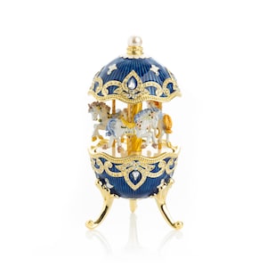 Puede incluir: Una caja de música de carrusel decorativa azul y dorada. La caja en forma de huevo presenta un carrusel con varios caballos blancos y beige. El carrusel está rematado con una perla y está sostenido por patas doradas. La caja está adornada con pedrería.