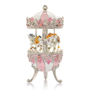 May include: A decorative carousel music box in pink, white, and silver. The carousel features three white horses with colorful manes and tails. The top and base are adorned with ornate detailing and pink accents. The music box stands on three silver legs.