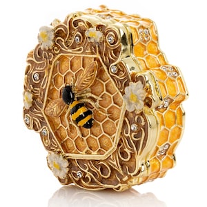 May include: A decorative, gold-colored trinket box shaped like a honeycomb. The box features a detailed bee and honeycomb design, with small white flowers and sparkling accents. The box is likely made of metal and has a glossy finish.