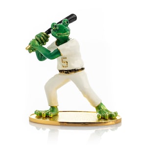 May include: A green frog figurine wearing a white baseball uniform with the number "5" on the front. The frog is holding a black baseball bat and is standing on a gold base.