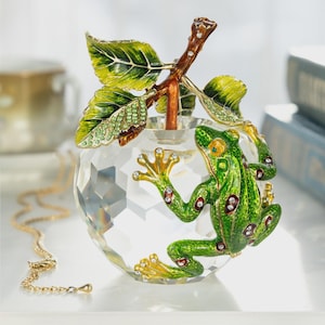 May include: A decorative glass orb with a green frog figurine. The frog has red and white accents and is perched on the orb. The orb has a brown stem with green leaves. A gold necklace is in the foreground.