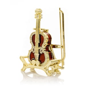 May include: A gold-coloured miniature violin figurine with a dark red body and clear crystal accents. The violin is set on a matching gold stand with a bow. The figurine is a decorative item.