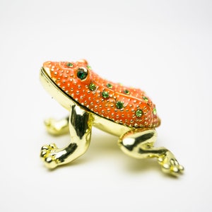 May include: A decorative frog figurine with an orange body and gold-colored legs. The frog's body is adorned with small green gemstones and gold accents. The figurine is a unique decorative item.