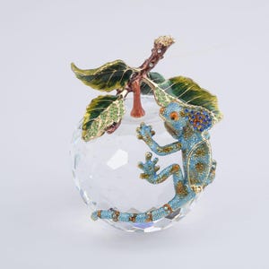 May include: A decorative crystal apple with a blue and gold lizard climbing on it. The lizard has orange eyes and is detailed with blue and gold accents. The apple has green and gold leaves and a brown stem.