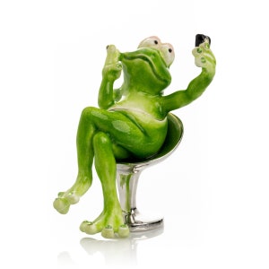 May include: A green ceramic frog figurine sitting on a silver chair, holding a black cell phone and taking a selfie.