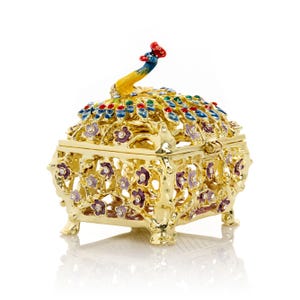 May include: A gold-colored, ornate jewelry box with a peacock design. The box features a hinged lid, floral cutouts, and is embellished with colorful jewels. The peacock's head and tail are prominently displayed on top of the box.