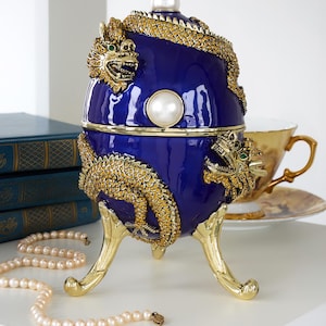 Puede incluir: Una caja decorativa azul cobalto en forma de huevo con adornos de dragón dorados y un acento de perla. La caja se apoya en patas doradas y presenta un cierre de perla. Un collar de perlas y una taza de té están en el fondo.