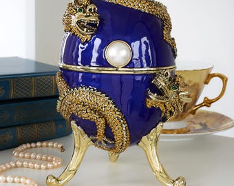 Puede incluir: Una caja decorativa azul cobalto en forma de huevo con adornos de dragón dorados y un acento de perla. La caja se apoya en patas doradas y presenta un cierre de perla. Un collar de perlas y una taza de té están en el fondo.