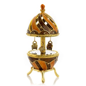 May include: A gold-tone carousel music box with a brown and orange striped egg-shaped base. The carousel has three golden seats and a gold-tone roof with a brown and orange striped design.