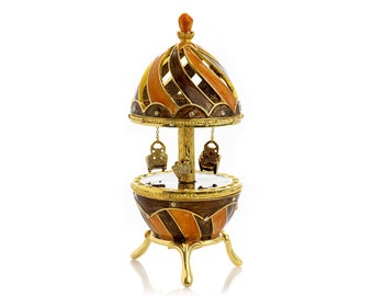 May include: A gold-tone carousel music box with a brown and orange striped egg-shaped base. The carousel has three golden seats and a gold-tone roof with a brown and orange striped design.