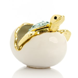 Może przedstawiać: Ozdobne pudełko na drobiazgi w kształcie żółwia wychodzącego z jaja. Żółw jest złoty z turkusowym pancerzem, a jajko jest białe ze złotymi zdobieniami. Pudełko jest przeznaczone do przechowywania małych przedmiotów.