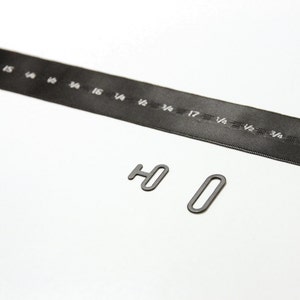 May include: Two black metal hardware pieces, one shaped like the letter "H" and the other shaped like a circle, on a white surface. A black ribbon with white markings is in the background.