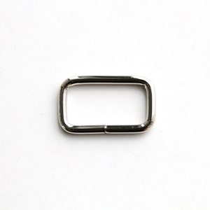 Puede incluir: Un anillo rectangular de metal plateado con bordes redondeados.