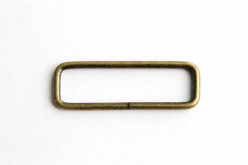 May include: A gold-toned metal rectangle ring, also known as a D-ring, with a smooth finish. This ring is commonly used for attaching straps, chains, or other accessories.