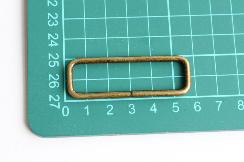 Peut inclure: Une boucle rectangulaire en m&eacute;tal de couleur dor&eacute;e avec des bords arrondis. La boucle est sur un tapis de coupe vert avec des chiffres blancs.