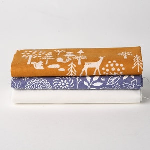 May include: A stack of three fabric squares with white woodland animal designs on a mustard yellow, a light blue, and a white background.
