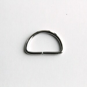 Puede incluir: Un anillo en D de metal plateado, un tipo de herraje utilizado para sujetar correas, cadenas u otros accesorios.