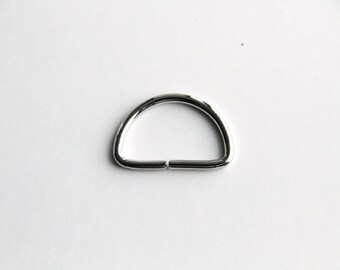 Puede incluir: Un anillo en D de metal plateado, un tipo de herraje utilizado para sujetar correas, cadenas u otros accesorios.