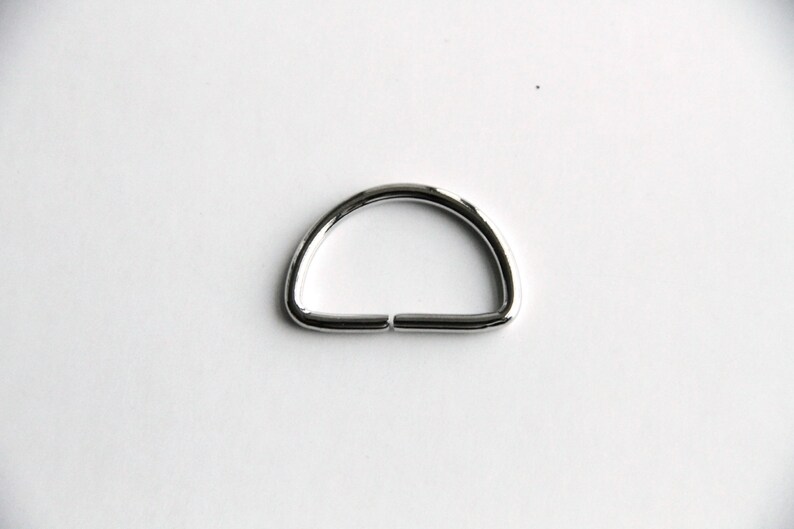 Puede incluir: Un anillo en D de metal plateado, un tipo de herraje utilizado para sujetar correas, cadenas u otros accesorios.
