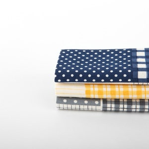 May include: A stack of four fabric squares with different patterns. The top square is navy blue with white polka dots, the second square is yellow with white and blue checked pattern, the third square is grey with white polka dots, and the bottom square is white with blue and grey checked pattern.