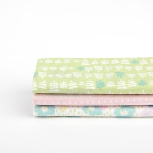 May include: A stack of three fabric squares with different patterns. The top square is green with a white leaf pattern, the middle square is pink with a white dot pattern, and the bottom square is white with a blue and yellow flower pattern.