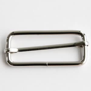 May include: A shiny, silver-colored rectangular slider buckle with a single bar across the middle. The buckle is made of metal and has rounded corners. This hardware component is used in various crafting and sewing projects.