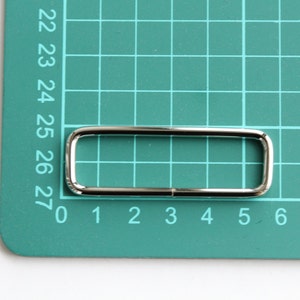 May include: A silver metal rectangular buckle with rounded corners, measuring approximately 2.5 inches in length and 0.5 inches in width.
