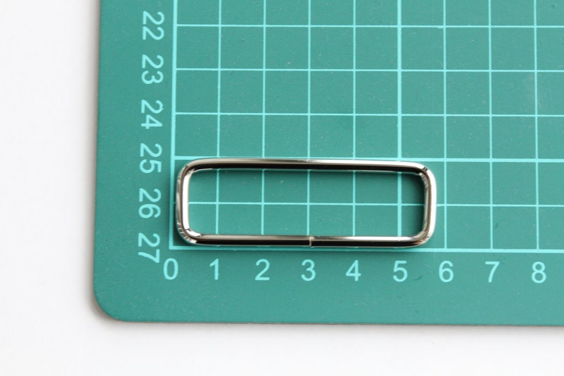 May include: A silver metal rectangular buckle with rounded corners, measuring approximately 2.5 inches in length and 0.5 inches in width.