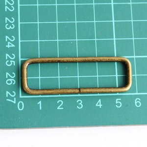 Peut inclure: Un anneau rectangulaire en m&eacute;tal dor&eacute;, &eacute;galement appel&eacute; anneau en D, est pr&eacute;sent&eacute; sur un tapis de coupe vert. L'anneau mesure environ 3 cm de long et 1 cm de large.