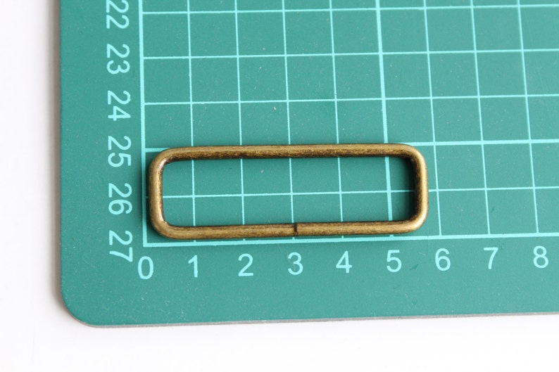 Peut inclure: Un anneau rectangulaire en m&eacute;tal dor&eacute;, &eacute;galement appel&eacute; anneau en D, est pr&eacute;sent&eacute; sur un tapis de coupe vert. L'anneau mesure environ 3 cm de long et 1 cm de large.