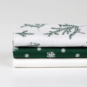 Op de afbeelding: Drie gevouwen stoffen servetten met een witte achtergrond en groene en witte patronen. De bovenste servet heeft een groen bloemmotief, de middelste servet heeft een groen sneeuwvlokmotief en de onderste servet is effen wit.