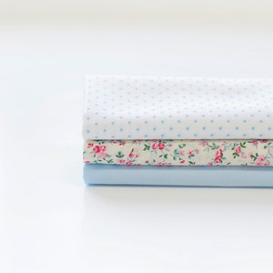 May include: A stack of three fabric squares in shades of blue and white. The top square has a white background with small blue polka dots. The middle square has a white background with a floral pattern in pink and green. The bottom square is solid light blue.
