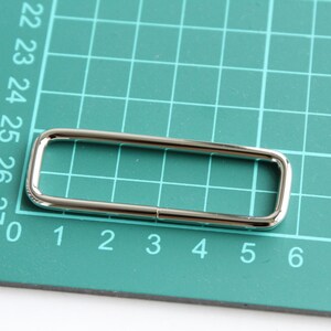 May include: A silver metal D-ring with a rectangular shape, measuring approximately 2.5 inches in length and 1 inch in width. The ring is lying on a green cutting mat with numbered grid lines.
