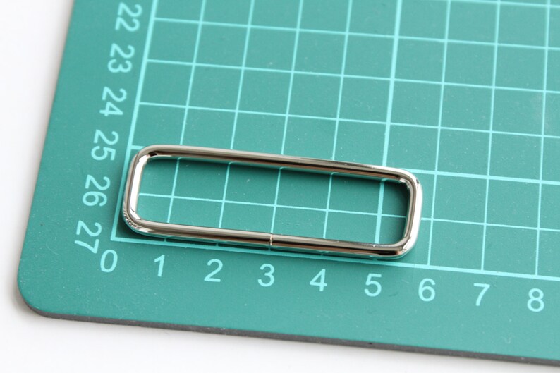 May include: A silver metal D-ring with a rectangular shape, measuring approximately 2.5 inches in length and 1 inch in width. The ring is lying on a green cutting mat with numbered grid lines.
