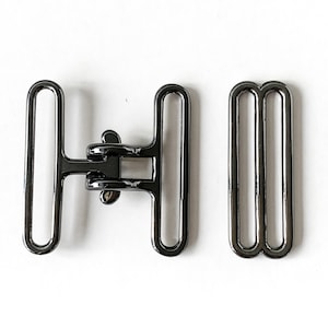 Puede incluir: Una hebilla de metal negro con cierre deslizante y una correa de metal negro a juego. La hebilla tiene un diseño de doble bucle.