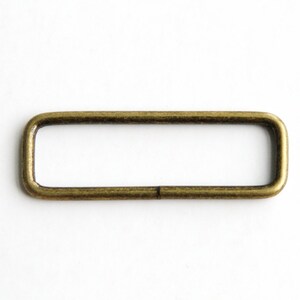 Puede incluir: Un anillo rectangular de metal dorado, también conocido como anillo en D, con un acabado liso. Este anillo se utiliza comúnmente para sujetar correas, cadenas u otros herrajes a bolsos, cinturones u otros accesorios.