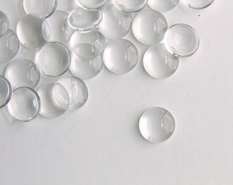 May include: A group of clear glass cabochons, also known as glass domes, scattered on a white surface. These glass domes are used for jewelry making, crafting, and other decorative purposes.