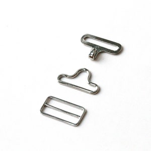 May include: Three silver metal hardware pieces for sewing or crafting projects. The pieces include a rectangular buckle, a D-ring, and a loop.