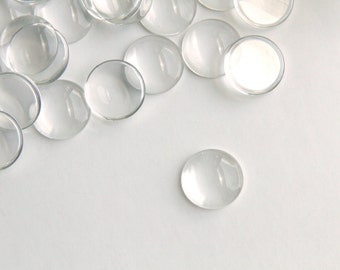 May include: A group of clear glass cabochons, some are stacked on top of each other, on a white surface. One cabochon is separated from the group.